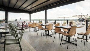 Een terras met tafels en stoelen, grote ramen en uitzicht op het water.