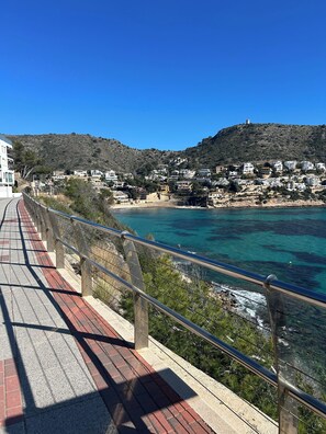 A coastal walkway with a metal railing, leading to a seaside town with buildings on a hillside. - Modern Studio apartment in the center of Moraira (VC)