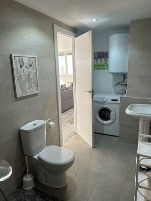 A modern bathroom with a toilet, bidet, and sink. A washing machine is visible in the background, and a painting is on the wall. - Modern Studio apartment in the center of Moraira (VC)