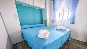 A single bed with a blue bedspread, a white headboard, and a window with blue curtains. - Sea View - 40 Parallelo Mobile Home (Camerota)