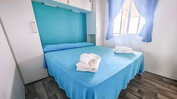 A single bed with a blue bedspread, a white headboard, and a window with blue curtains.