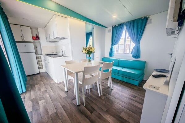Une cuisine compacte avec des meubles blancs, une table à manger avec des chaises, un canapé turquoise et une fenêtre avec des rideaux bleus.