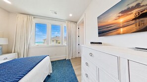 A hotel room with a bed, a dresser, a painting of a sunset, and a window with curtains. - 83 Dune Lane - Oceanfront Gem- Discover this NEW 7-Bedroom Haven (Hilton Head Island)