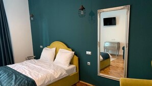 A room with a bed, a mirror, a yellow chair, and a wooden herringbone floor.