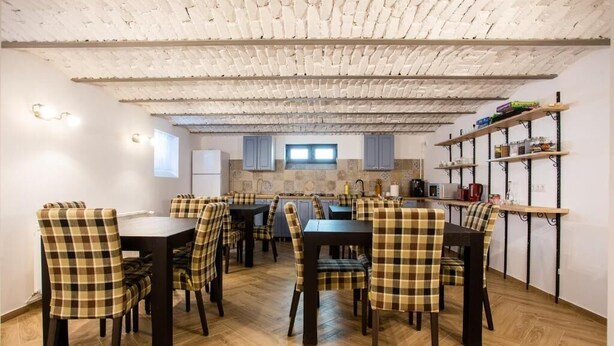 A dining area with checkered chairs, a wooden table, a kitchen in the background, and a brick ceiling.