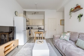 A modern kitchen with white cabinets, a dining area with a wooden table and chairs, a white sofa with patterned cushions, and a TV on a wooden stand. - Stylish 1 -Bed Leith Apartment | Near Shore & Tram (Edinburgh)
