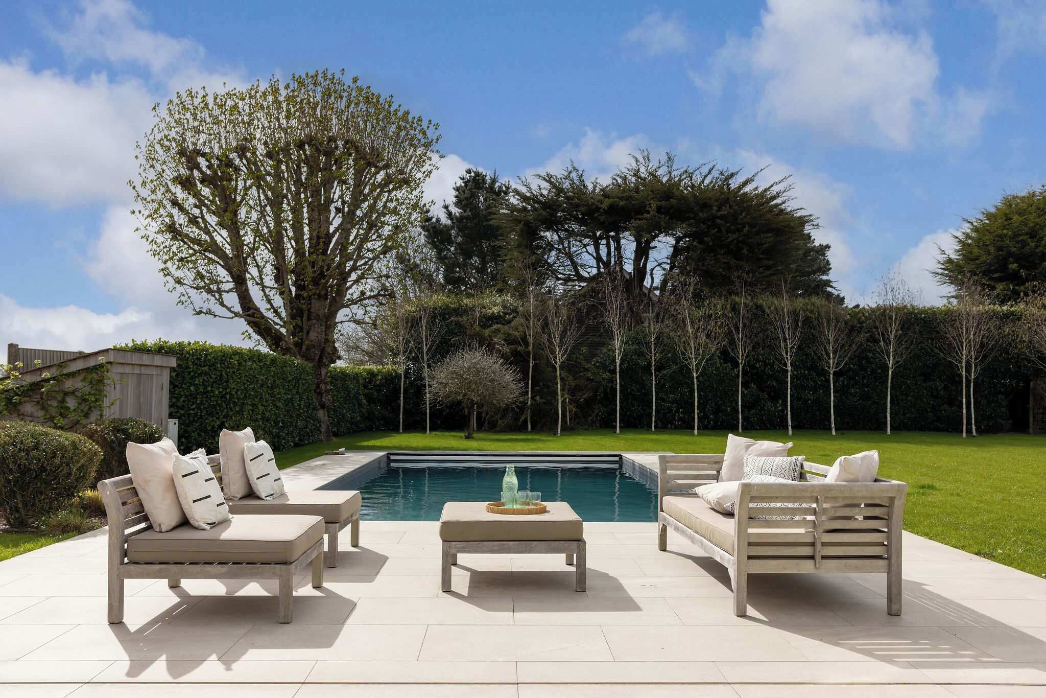 A patio with two sofas and a table, a swimming pool, and a hedge.