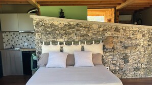 Una camera da letto con parete in pietra, travi in legno al soffitto, un letto con cuscini e una cucina sullo sfondo.