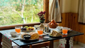 Mesa de café da manhã com frutas, pão e bebidas, um vaso com flores, e vista para o verde lá fora.