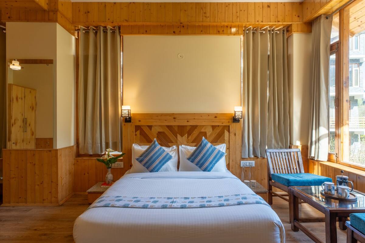 A wooden-paneled room with a bed, two bedside lamps, a chair, and a table with a cup and saucer.
