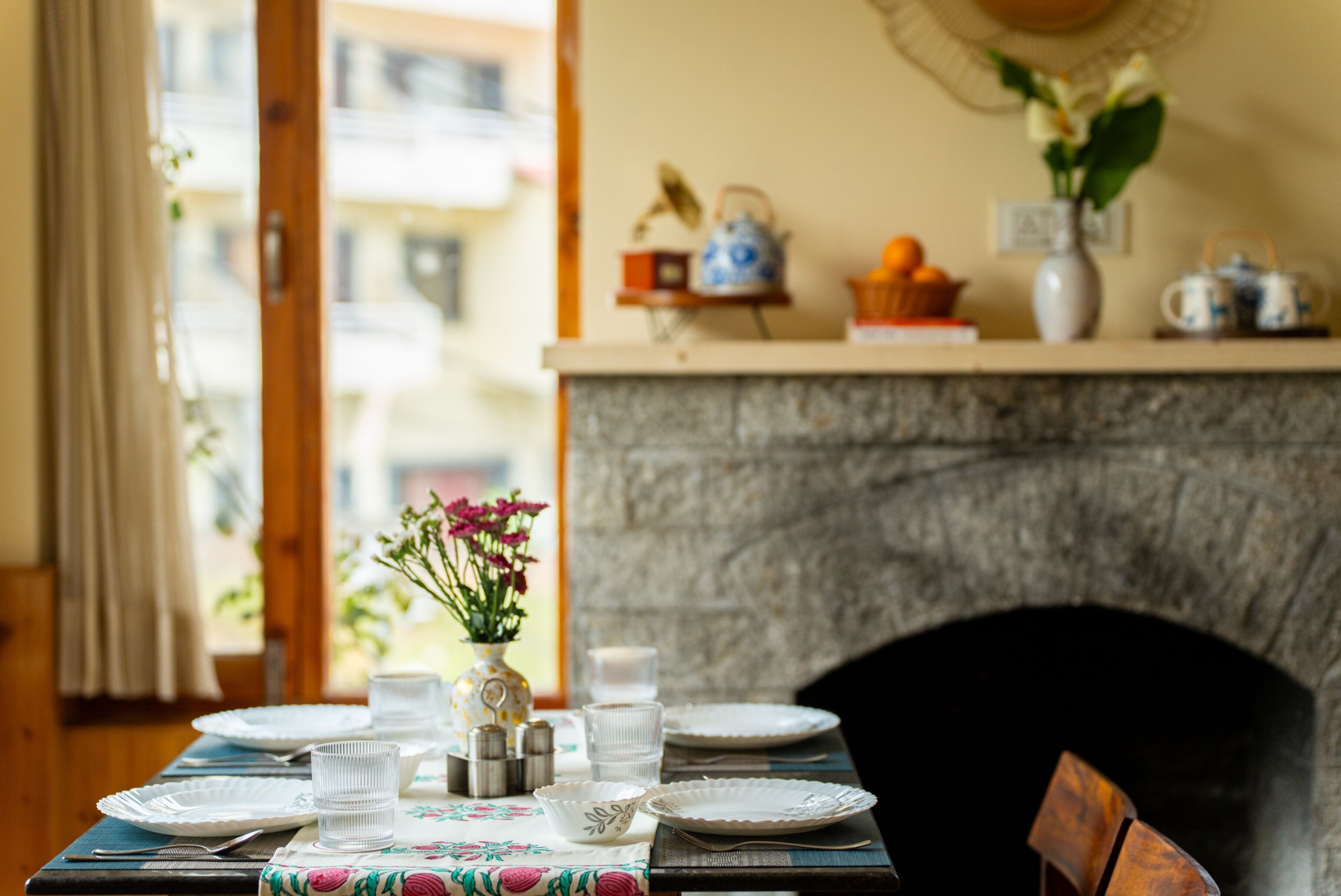 Ein Esstisch ist gedeckt mit Tellern, Gläsern und einer Blumenvase, im Hintergrund ist ein Kamin zu sehen.