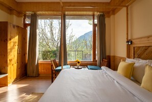 A wooden room with a large bed, a window with curtains, a wooden wardrobe, and a view of trees and mountains.