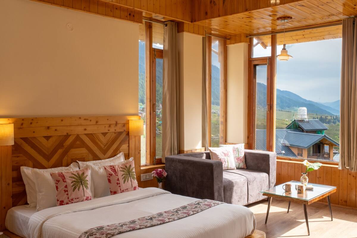 A wooden-paneled room with a bed, two armchairs, a small table, and large windows offering a mountain view.