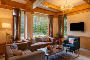 Una sala de estar acogedora con sofá, sillón y mesa de centro, un televisor instalado en la pared y ventanas grandes con cortinas.
