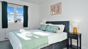 Een slaapkamer met een bed, kussens, een nachtkastje, een lamp, een raam met gordijnen en een aan de muur hangend schilderij.
