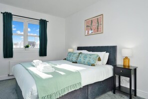 A bedroom with a bed, pillows, a bedside table, a lamp, a window with curtains, and a wall-mounted picture. - Buie`s Runway Retreat (Edinburgh)
