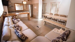 Una cocina moderna con vigas de madera, una zona de comedor con mesa y sillas de madera, y un salón con sofá y cojines.