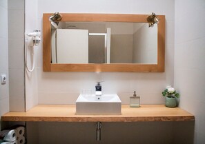 Une salle de bain avec un meuble-lavabo en bois, un lavabo blanc, un miroir, une douchette à main et un petit vase avec des fleurs.