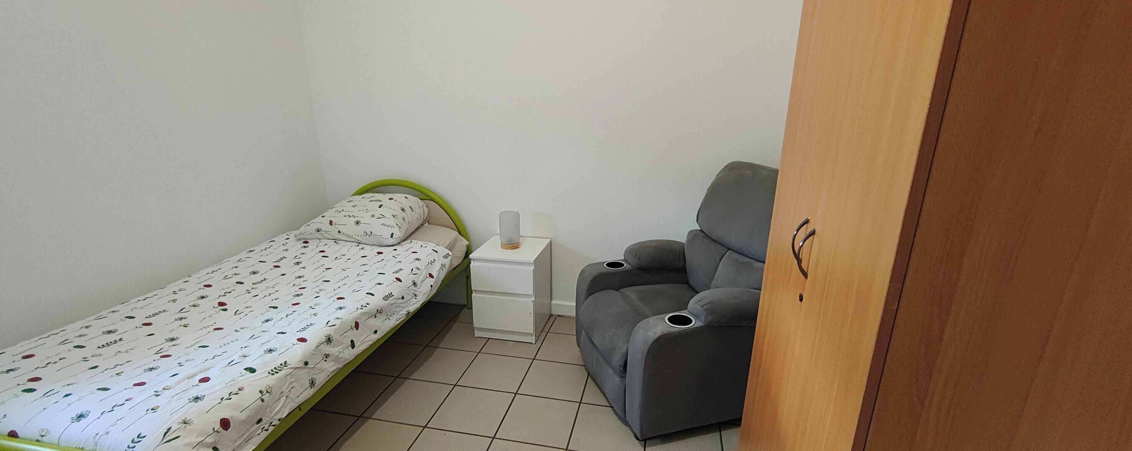 A small room with a single bed, a nightstand, a grey armchair, and a wooden wardrobe.