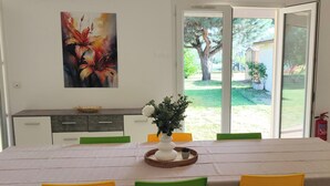 Un comedor con una mesa preparada para cuatro personas, un cuadro en la pared y vistas a un jardín a través de puertas de cristal.