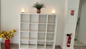 Una estantería blanca con una planta en maceta y velas encima, un jarrón con girasoles, y un extintor en el suelo.