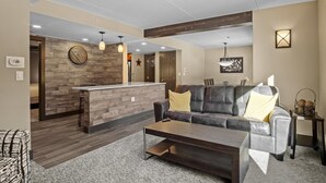 A modern living room with a dark leather sofa, a wooden coffee table, and a kitchen area with a wooden backsplash. - Slopeside Condo w/ Chairlift Views & Pool (Breckenridge)