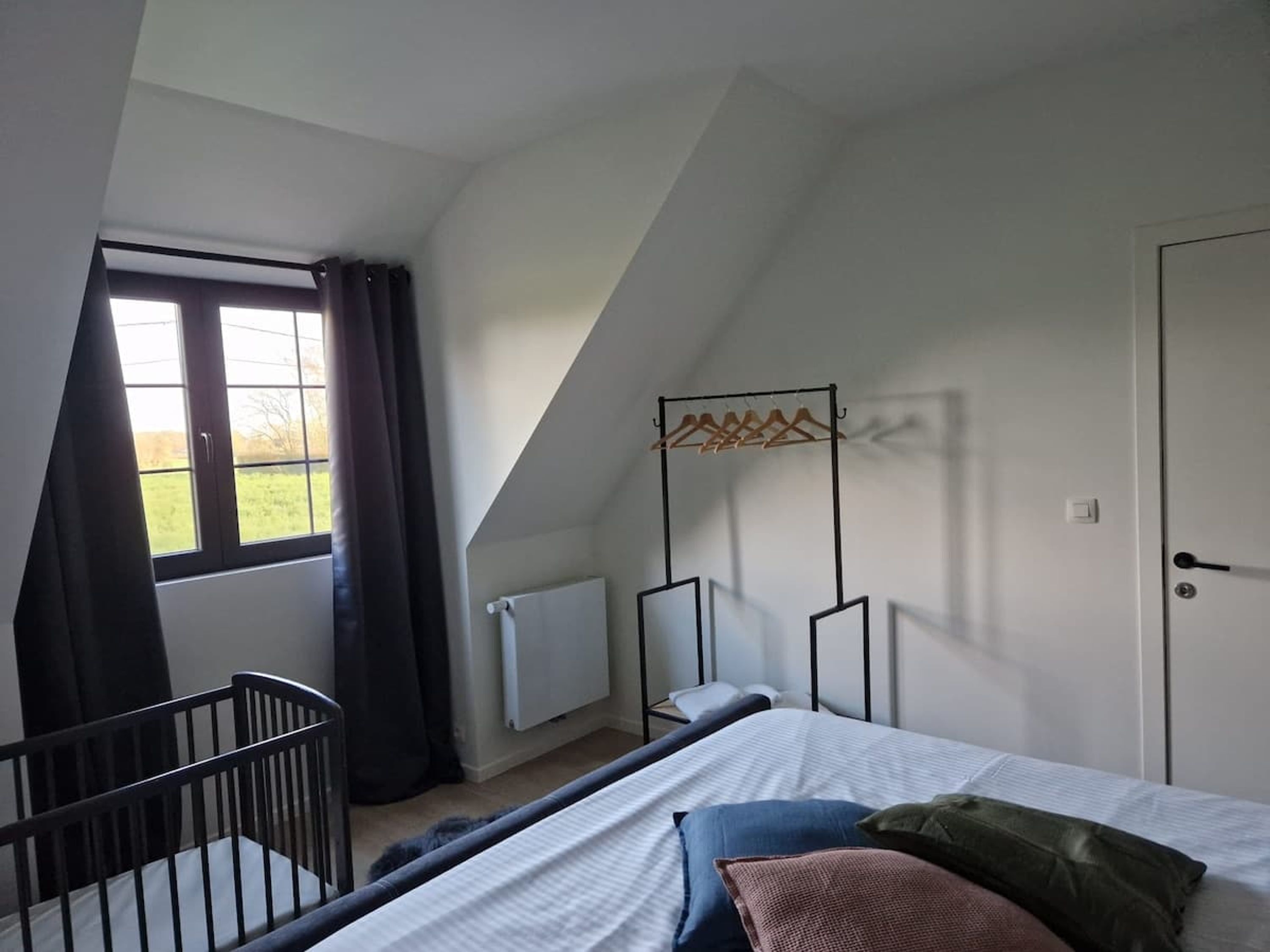 Ein Schlafzimmer mit schrägen Wänden, einem Fenster mit Vorhängen, einem Heizkörper, einem Bett mit Kissen, einem Babybett und einem Kleiderständer.