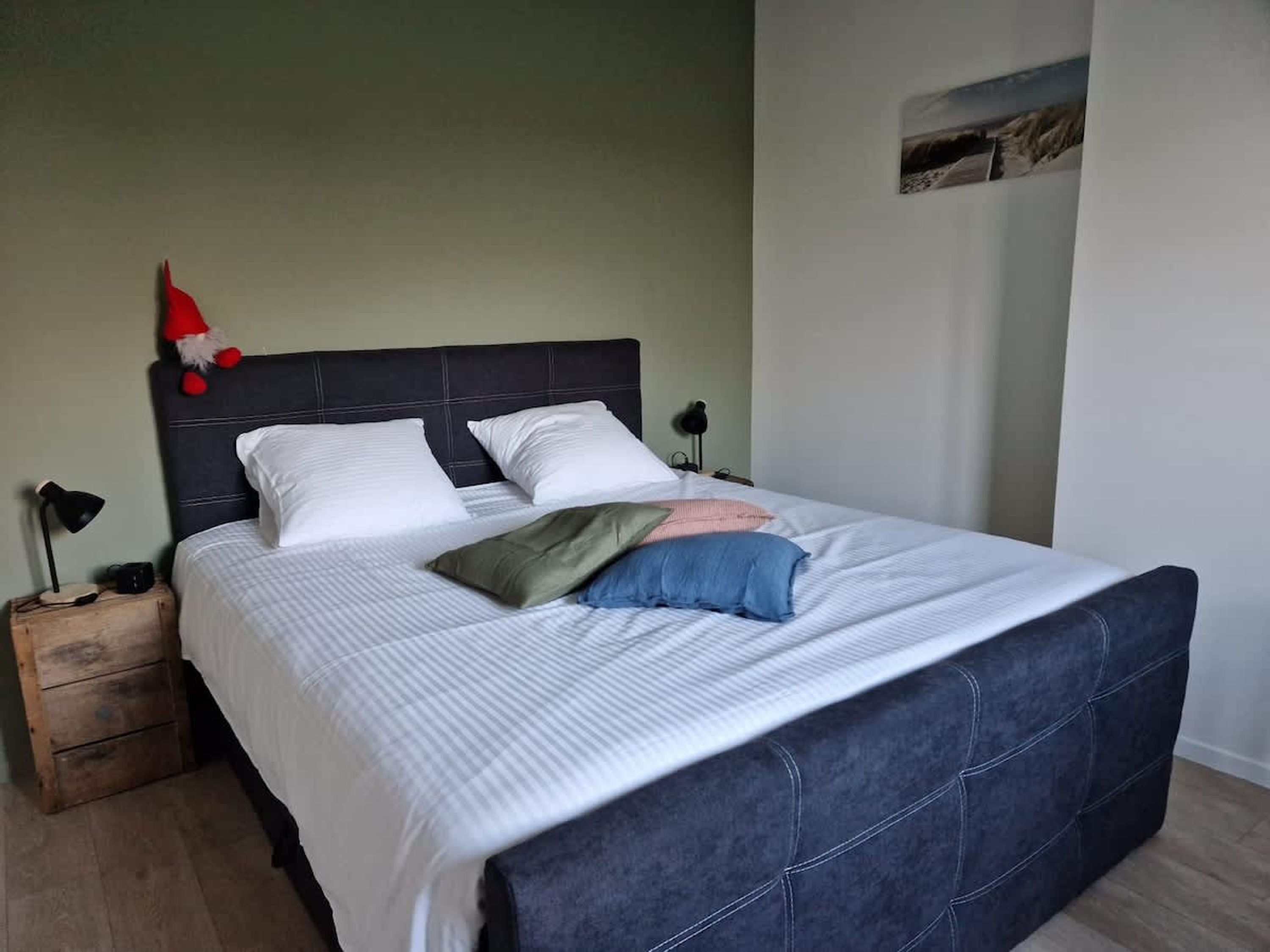 A neatly made bed with a dark headboard, a small wooden nightstand with a lamp, and a wall-mounted picture.