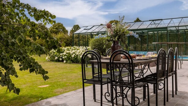 Un patio con tavolo e sedie, una serra sullo sfondo e un giardino con fiori e alberi.