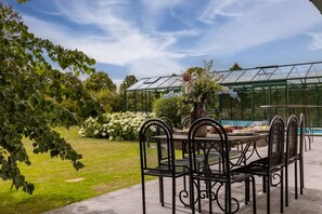 Un patio con tavolo e sedie, una serra sullo sfondo e un giardino con fiori e alberi.