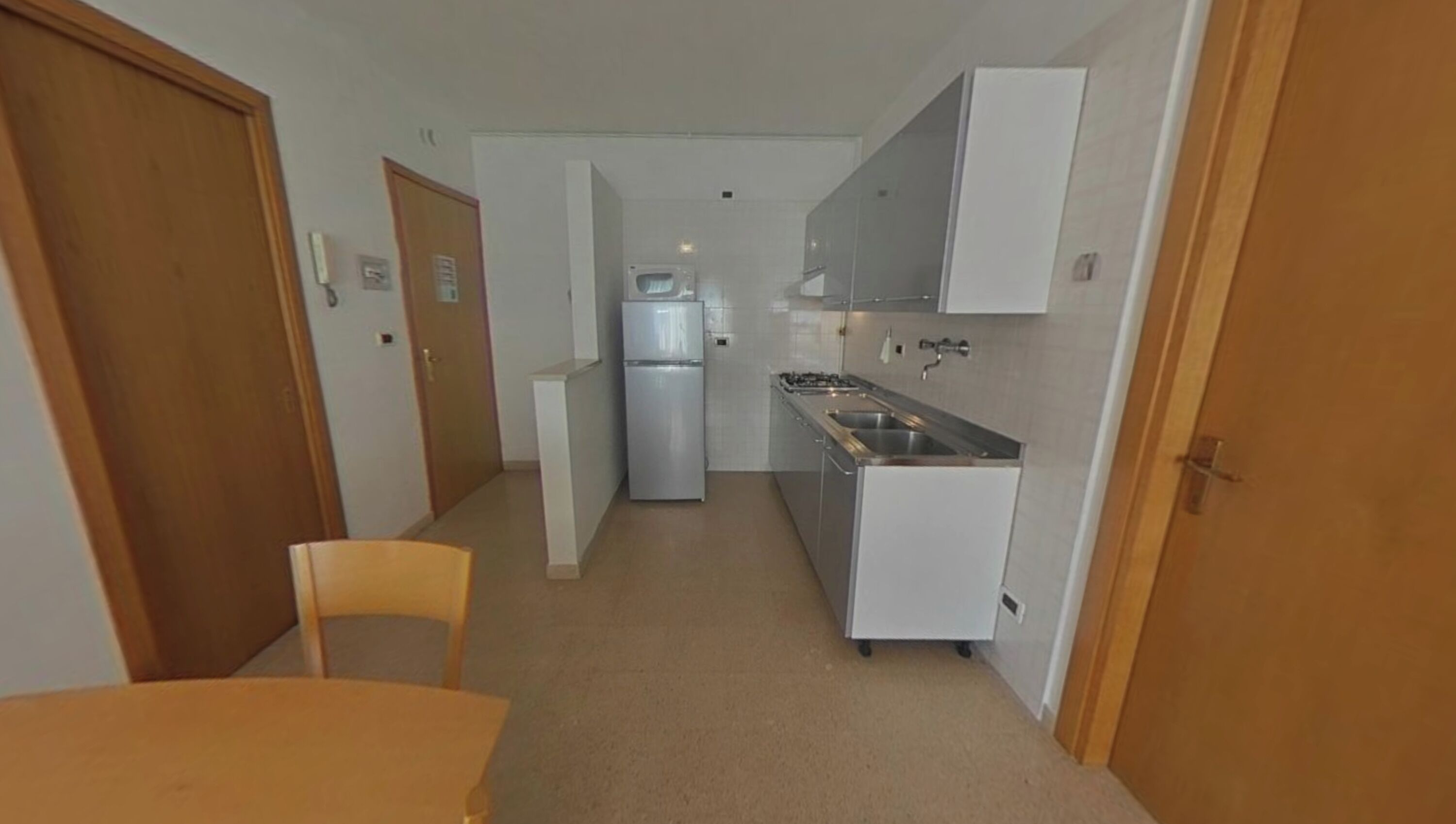 A narrow hallway leading to a kitchen with a refrigerator, stove, and sink. There is a wooden door on the left and a chair in the foreground.