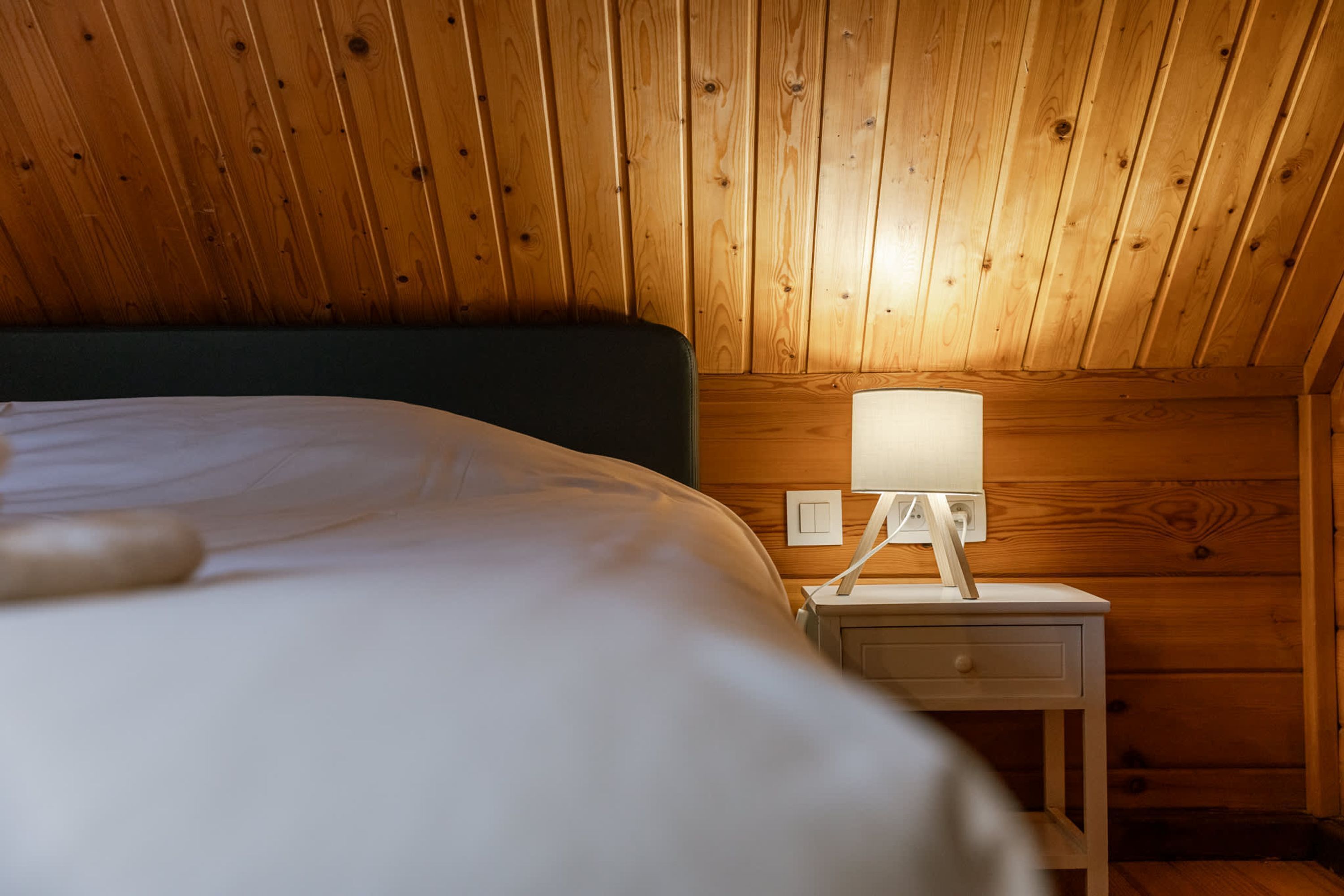 Une pièce aux murs lambrissés, comprenant un lit, une table de chevet avec une lampe et une prise de courant.