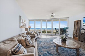 A living room with a large window, a ceiling fan, a sofa, a coffee table, and a view of the beach. - Caribe Resort 911D by Liquid Life (Orange Beach)
