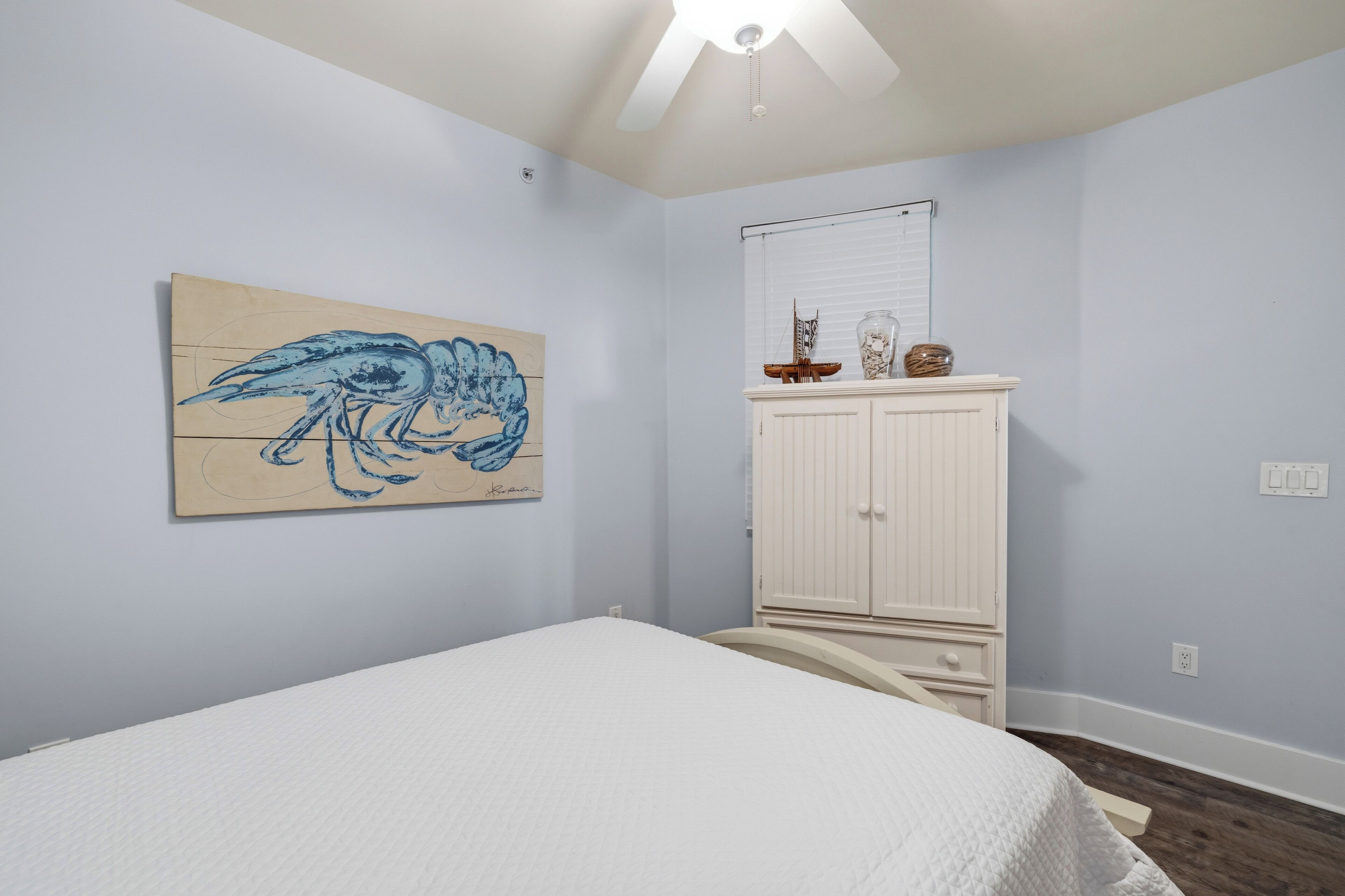 A bedroom with a bed, a white ceiling fan, a wall-mounted artwork of a bird, a white cabinet, and a window with blinds.