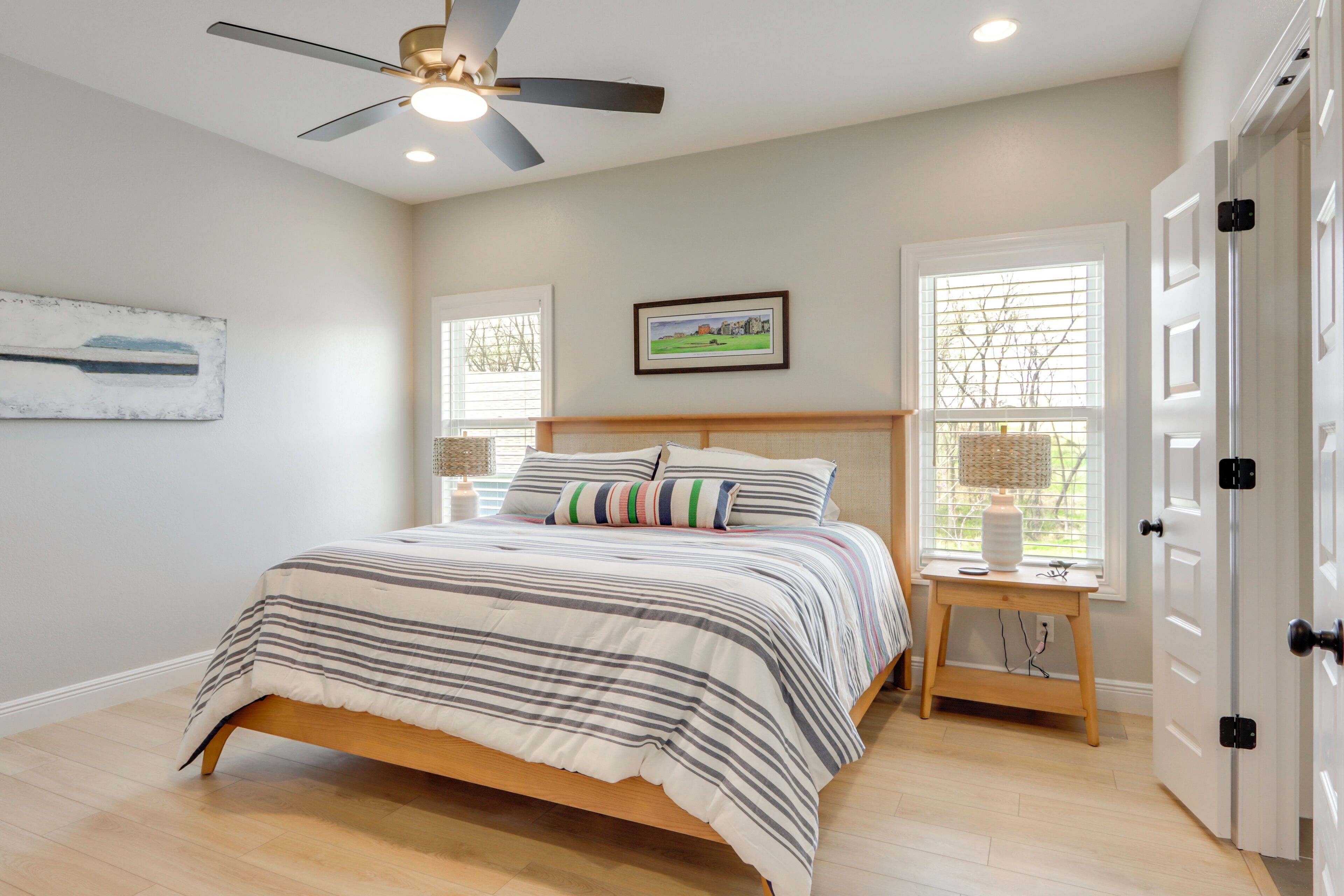 A bedroom with a bed, a ceiling fan, a window with blinds, a bedside table, and a door.
