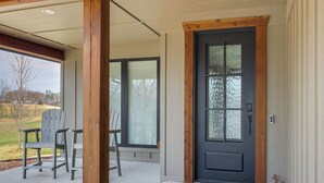 A covered porch with Adirondack chairs, a black door with glass panels, and a view of a grassy area. - Steps From the Water! Grand Lake Getaway w/ Patio (Afton)
