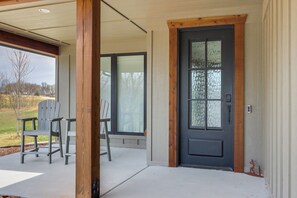 Un porche cubierto con sillas Adirondack, una puerta negra con paneles de vidrio y vista a un espacio cubierto de césped.