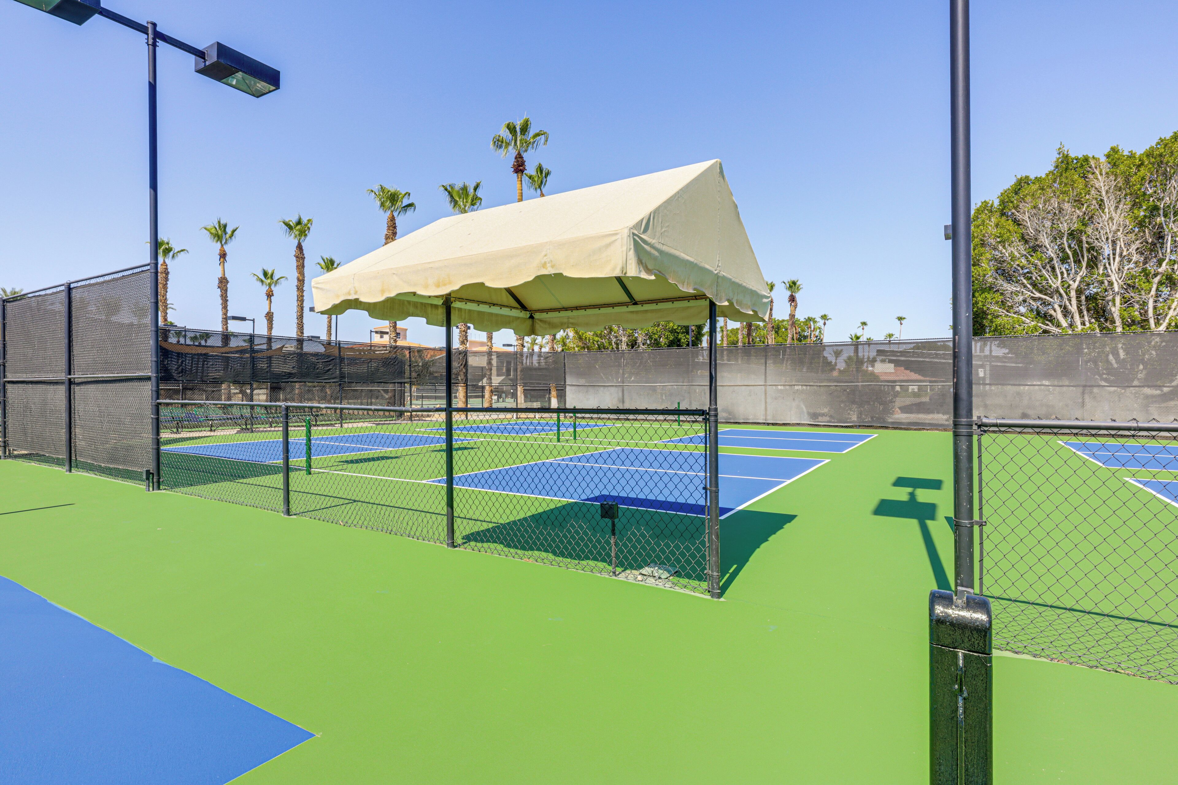 Cancha de tenis con una pérgola, cerca de malla y palmeras al fondo.