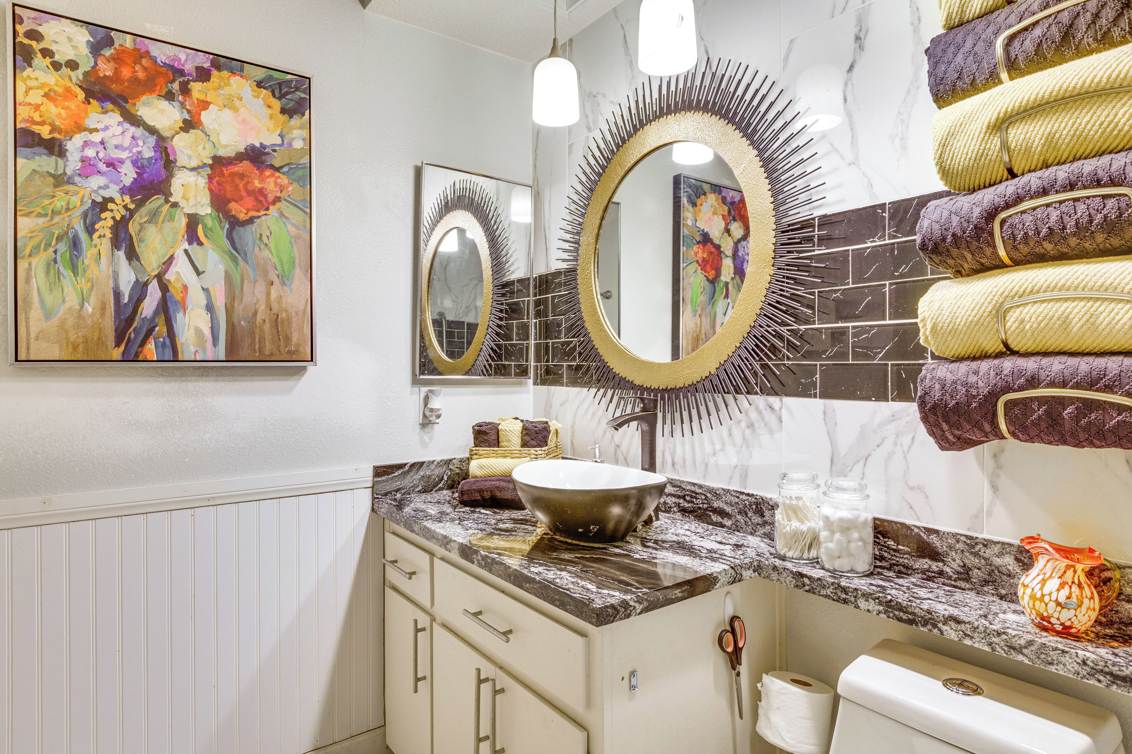 A bathroom with a marble countertop, a sink, a large mirror with a sunburst frame, a painting of flowers, and towels stacked on the wall.