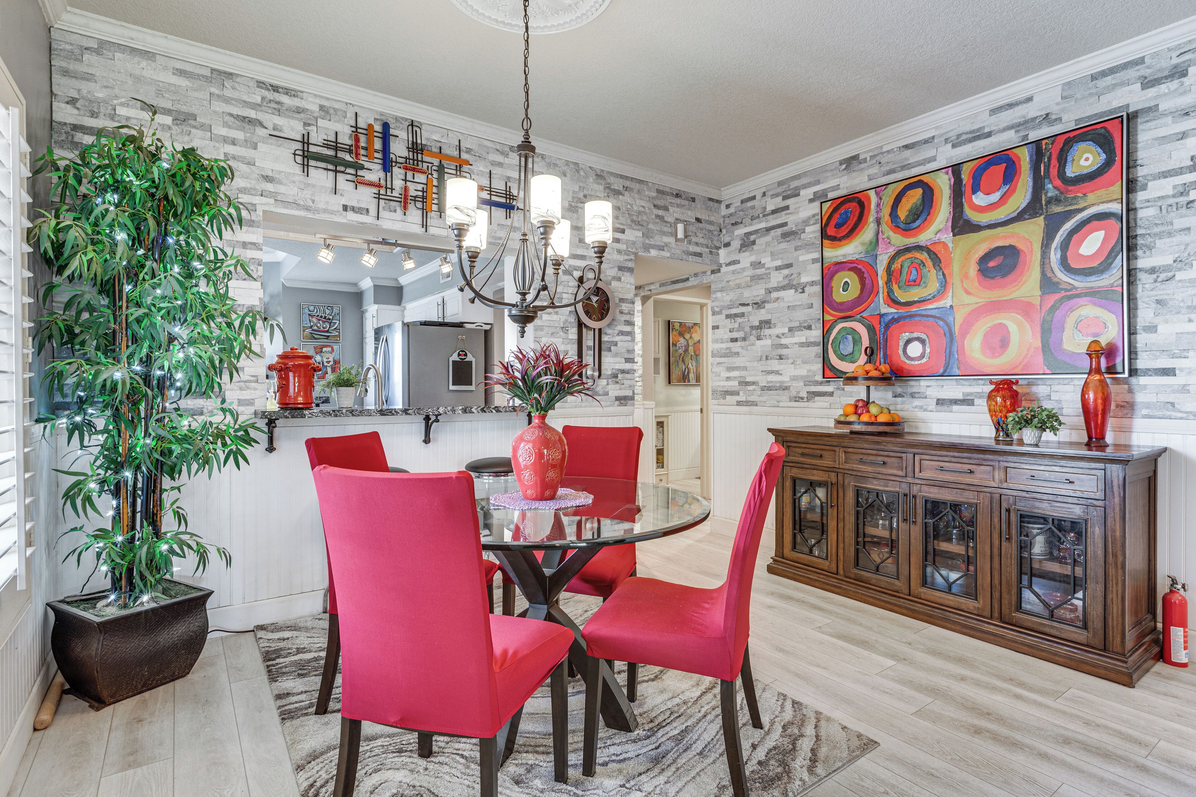 A modern kitchen with a dining area, featuring a round table, red chairs, a potted plant, and a colorful abstract painting.