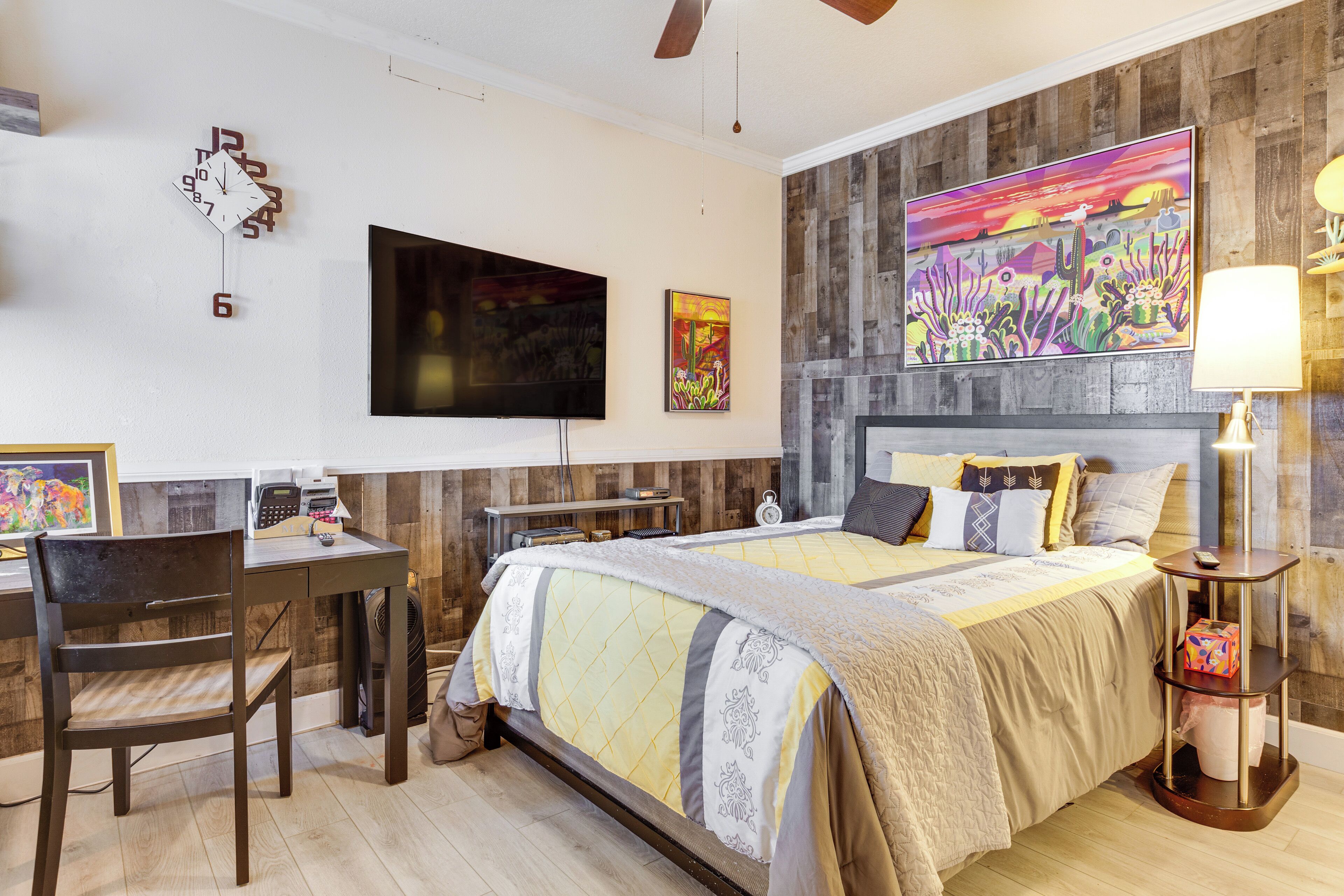 Een slaapkamer met een bed, een bureau, een stoel, een televisie en kunstwerken aan de wanden.
