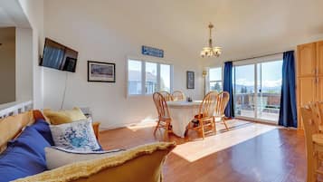 A spacious living area with a wooden floor, a sofa with blue cushions, a dining table with chairs, and a large window with a view of mountains.