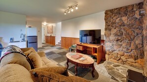 A living room with a stone wall, a television, a wooden desk, and a sofa. - < 1 Mi to Pioneer Ave: Homer Base w/ Fire Pit! (Homer)