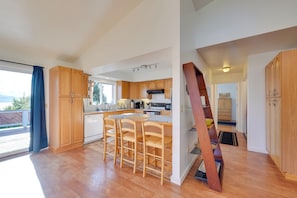 A spacious kitchen with wooden cabinets, a central island with bar stools, and a view of the outdoors through a large window. - < 1 Mi to Pioneer Ave: Homer Base w/ Fire Pit! (Homer)