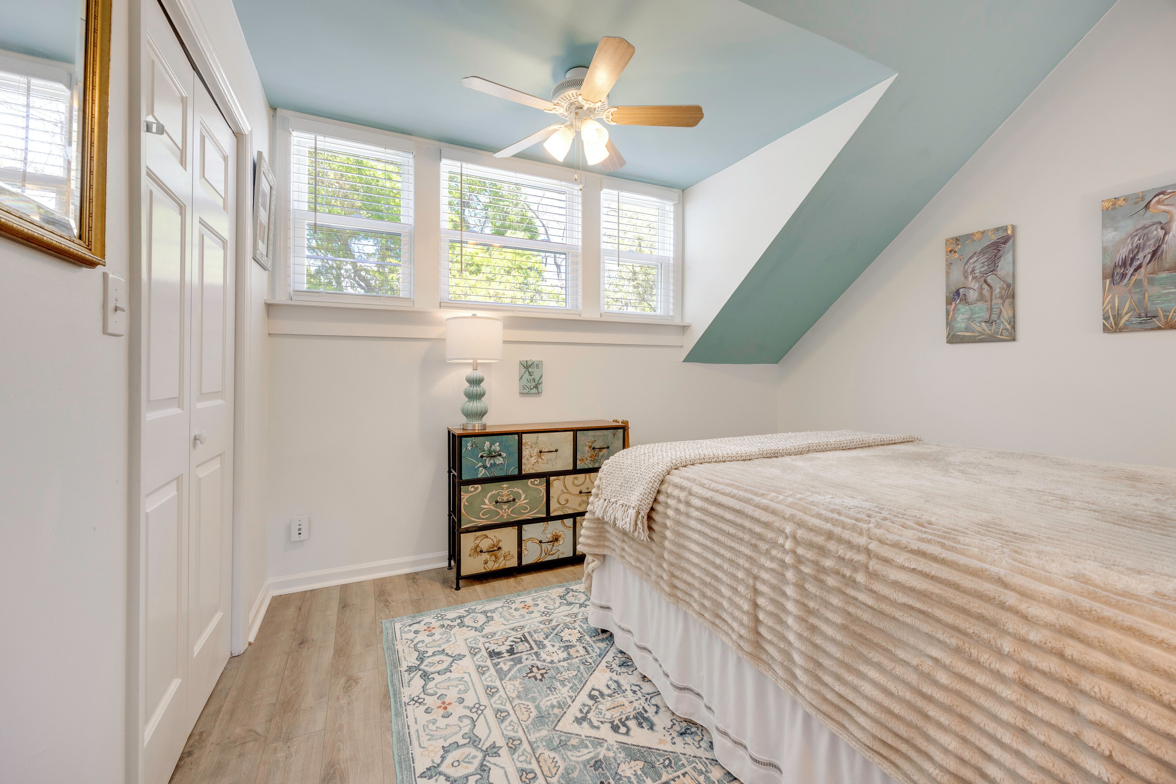 A bedroom with a sloped ceiling, a ceiling fan, a bed with a white bedspread, a nightstand with a lamp, and a wall-mounted shelf with decorative items.