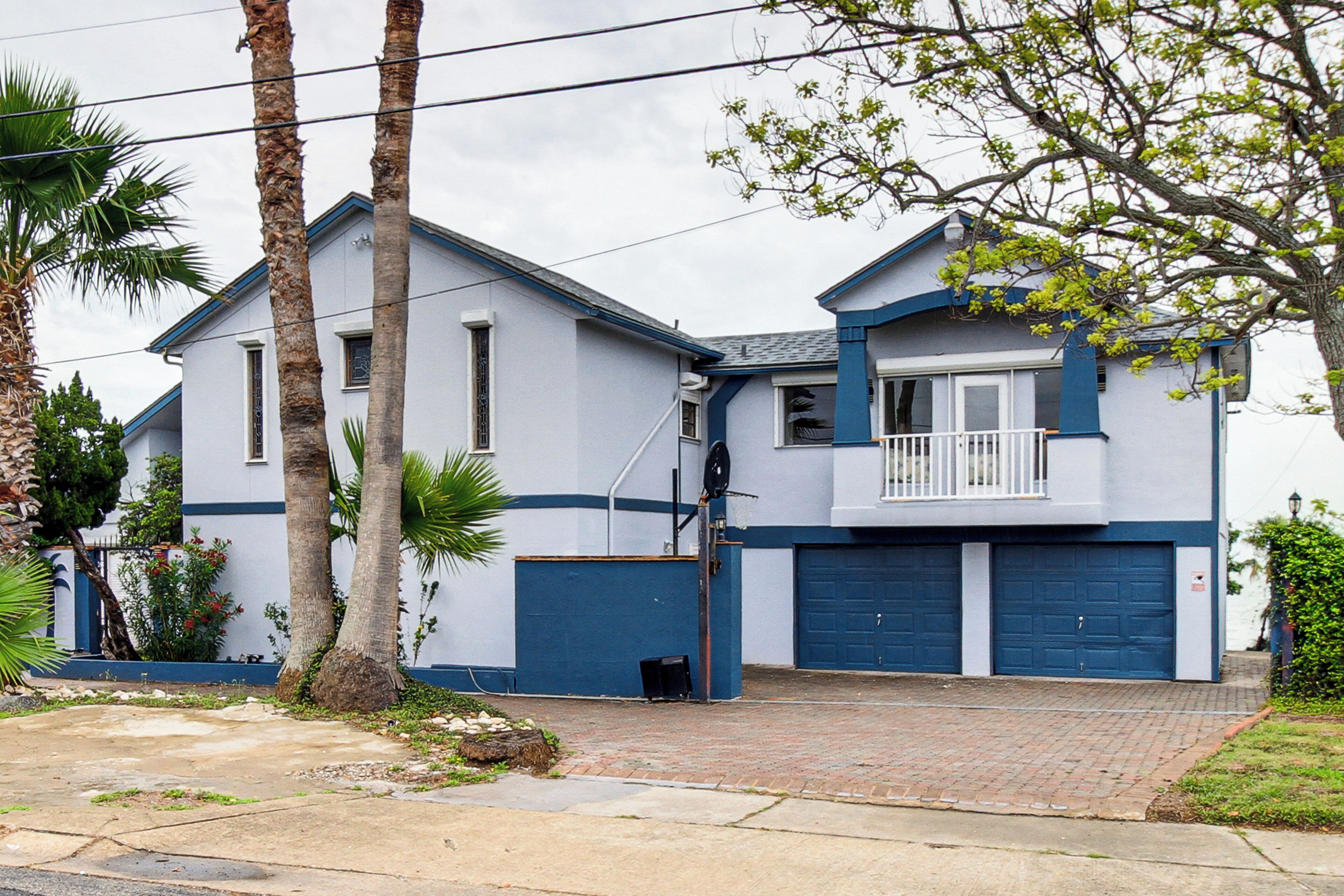A two-story house with a blue garage door, a balcony, and a small front yard with palm trees.