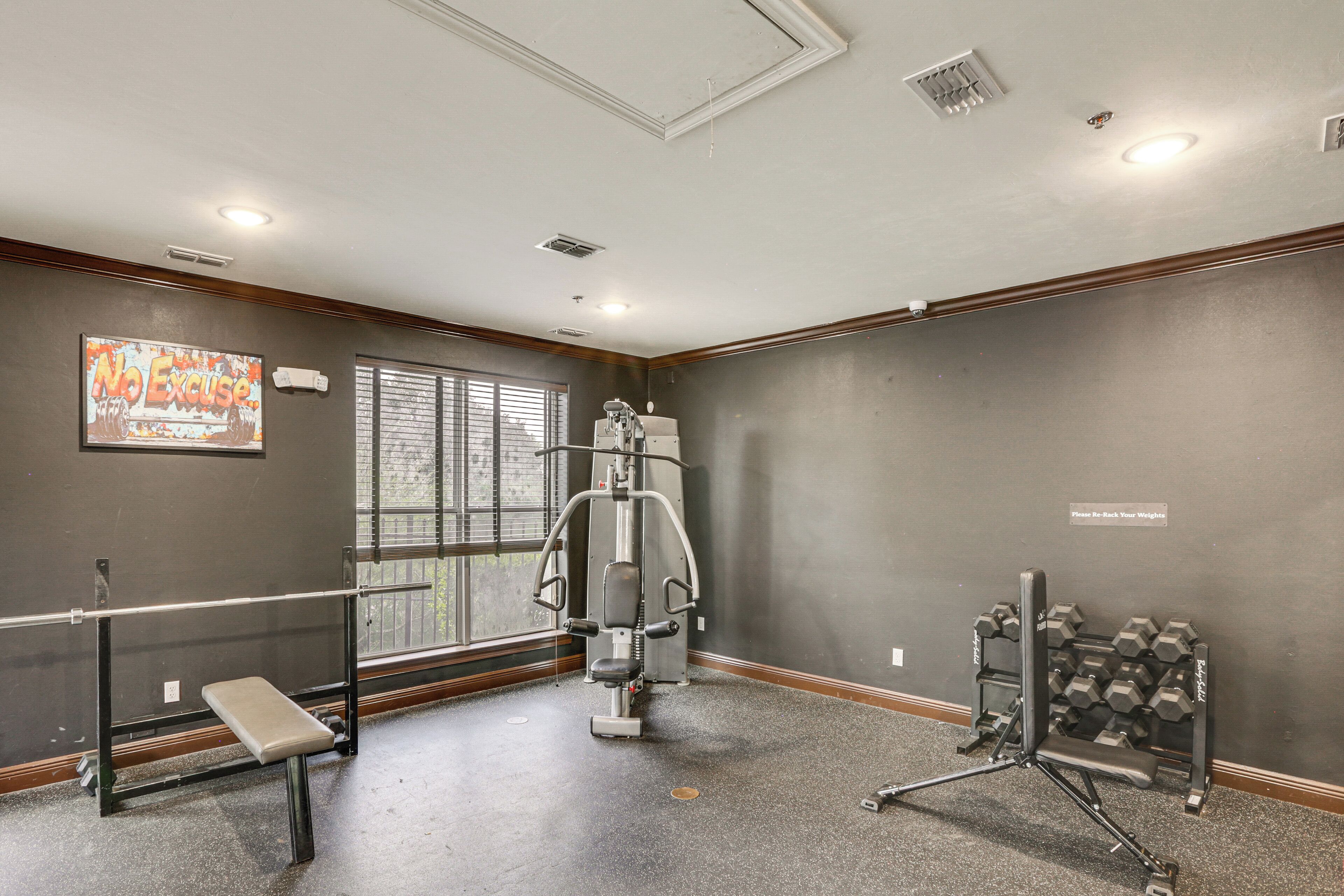 A gym with a bench, weight machine, dumbbells, and a window with blinds.