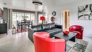 En moderne stue med en sort lædersofa, røde stole, et spiseområde med bord og stole, og et stort abstrakt billede på væggen.