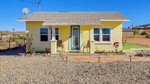 Une maison jaune à un seul étage, avec une porte bleue, deux fenêtres, une antenne parabolique et un petit jardin agrémenté de fleurs et d’une pelouse.