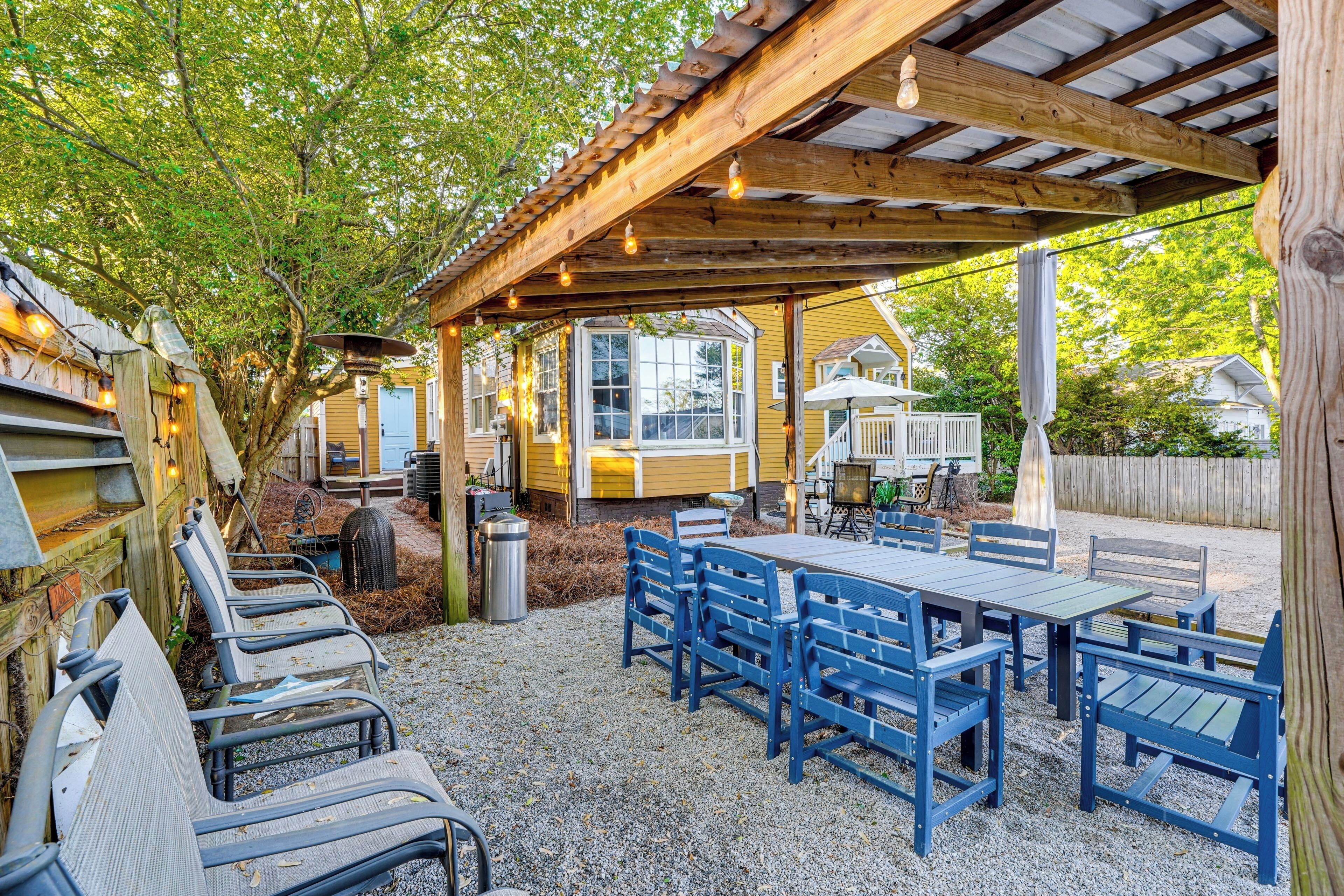 Un espace extérieur abrité, aménagé avec des tables et des chaises, une pergola illuminée de guirlandes lumineuses, et une vue sur une maison en arrière-plan.